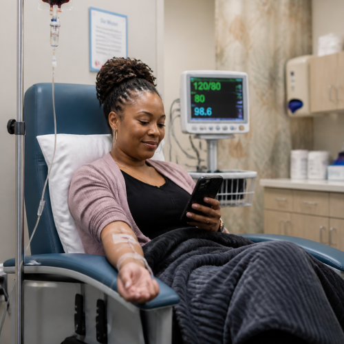 Woman donating blood in a clinic chair, IV line in her arm, monitoring screen with vitals in the background as she looks at her phone.